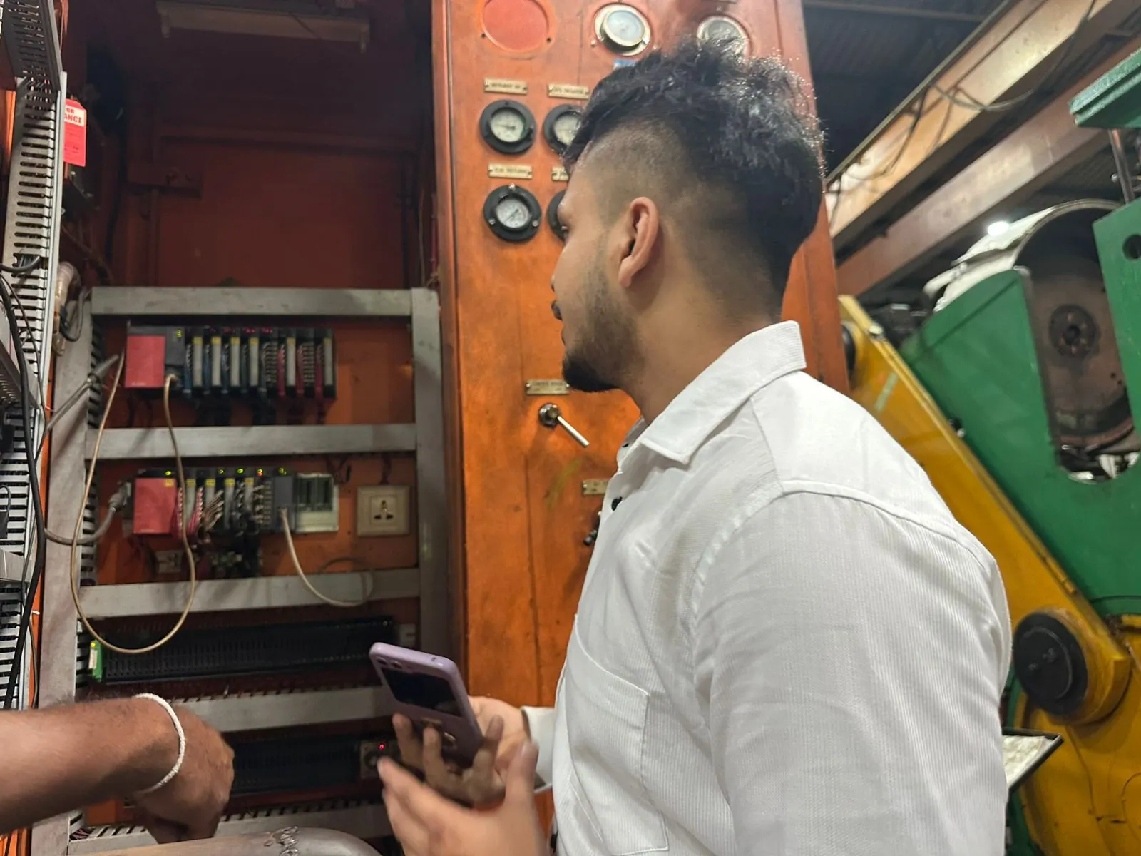 MechaCAD engineer examining an existing PLC control panel on a customer's factory floor during a site visit, with the machine's original gauge panel alongside